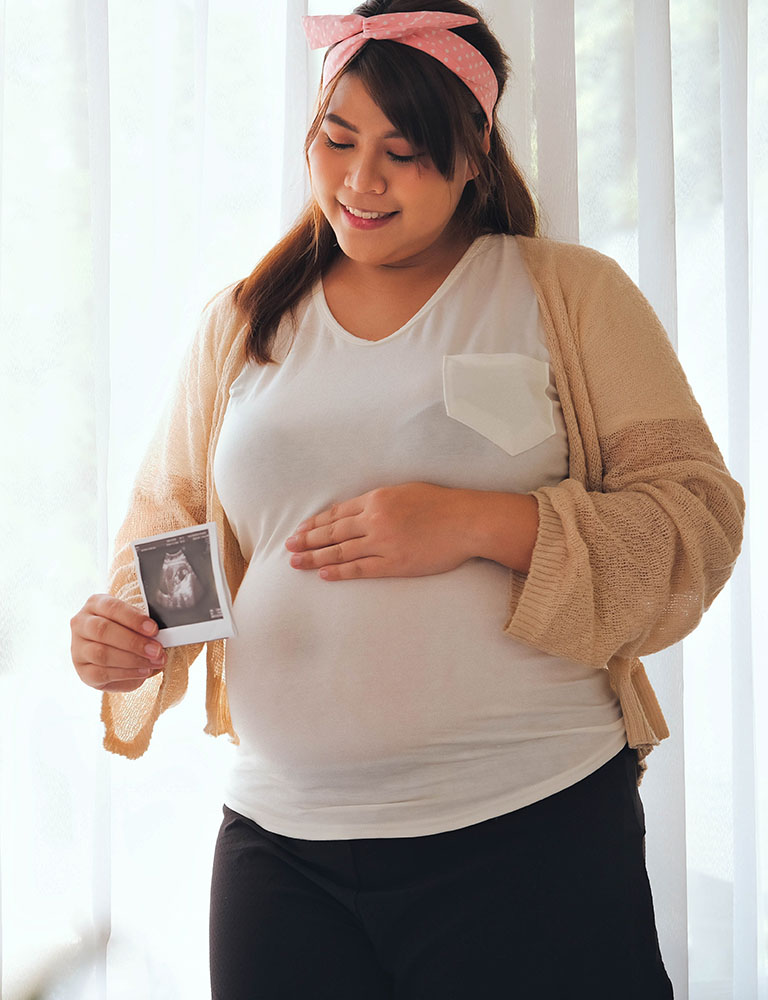 A pregnant woman smiles, holding an ultrasound photo in one hand and touching her belly with the other. She wears a light cardigan, white shirt, and a pink headband, standing in front of sheer curtains.