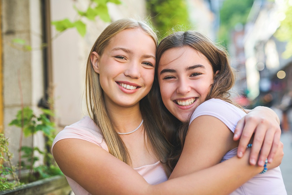 Two teenage girls smiling and hugging each other outdoors on a sunny day, with greenery and buildings in the blurred background. They appear happy and close, both wearing light-colored shirts.