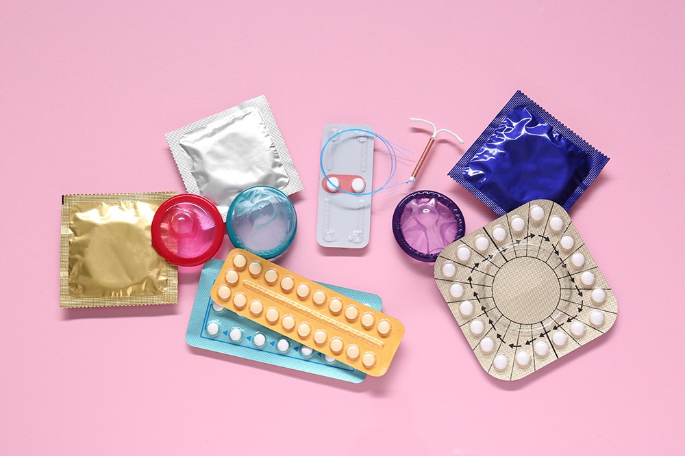A variety of birth control methods displayed on a pink background, including condoms, birth control pills, a contraceptive ring, and an intrauterine device (IUD).
