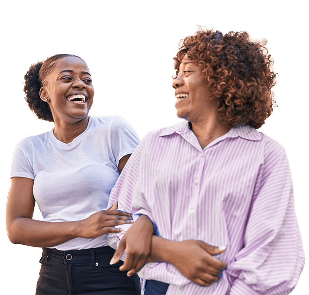 Two women smiling and laughing together, one wearing a white t-shirt and the other a light purple striped blouse, standing side by side against a plain, light background.