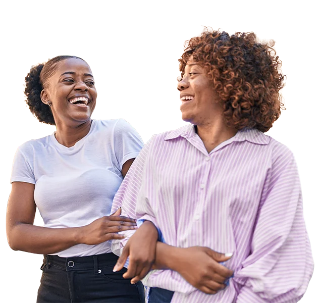 Two women smiling and laughing together, one wearing a white t-shirt and the other a light purple striped blouse, standing side by side against a plain, light background.