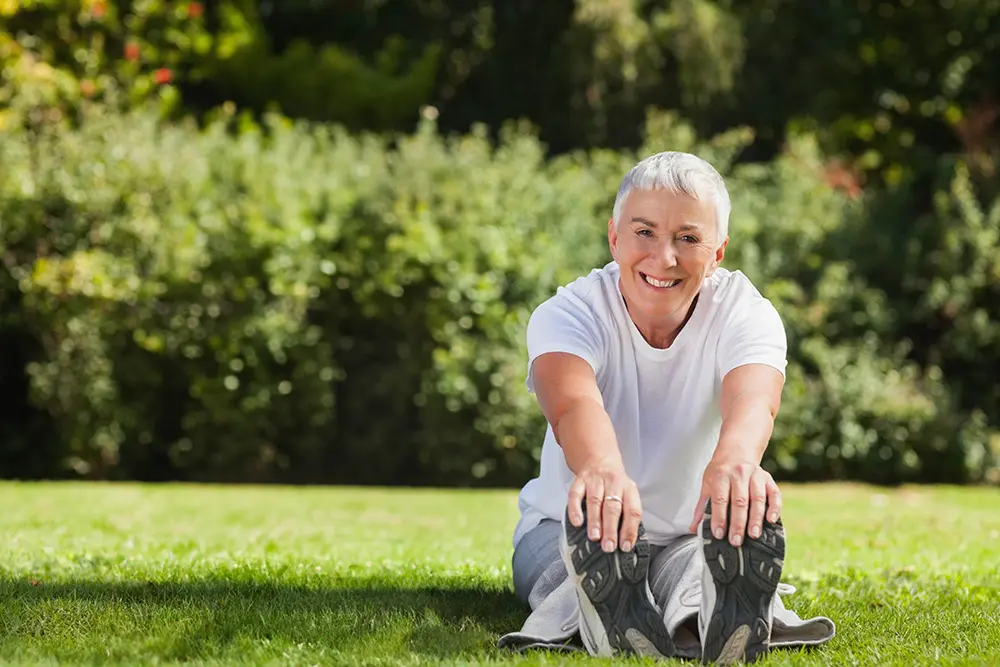 A smiling, active mature woman with short gray hair stretches toward her toes while sitting on green grass in a sunny park.