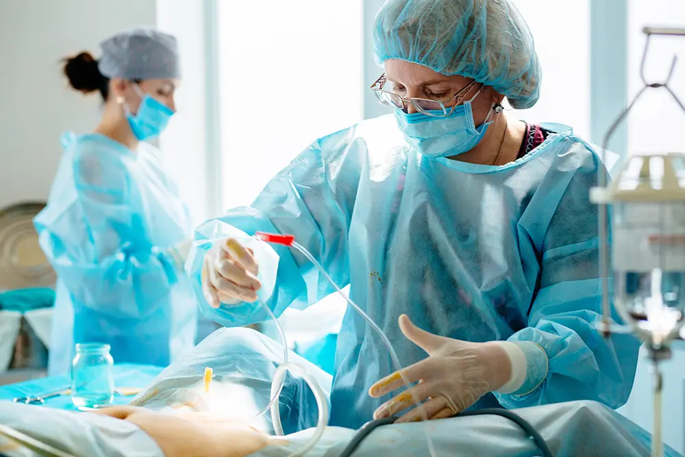 A surgeon in blue scrubs, mask, and gloves performs a medical procedure on a patient, while another medical professional works in the background in an operating room.