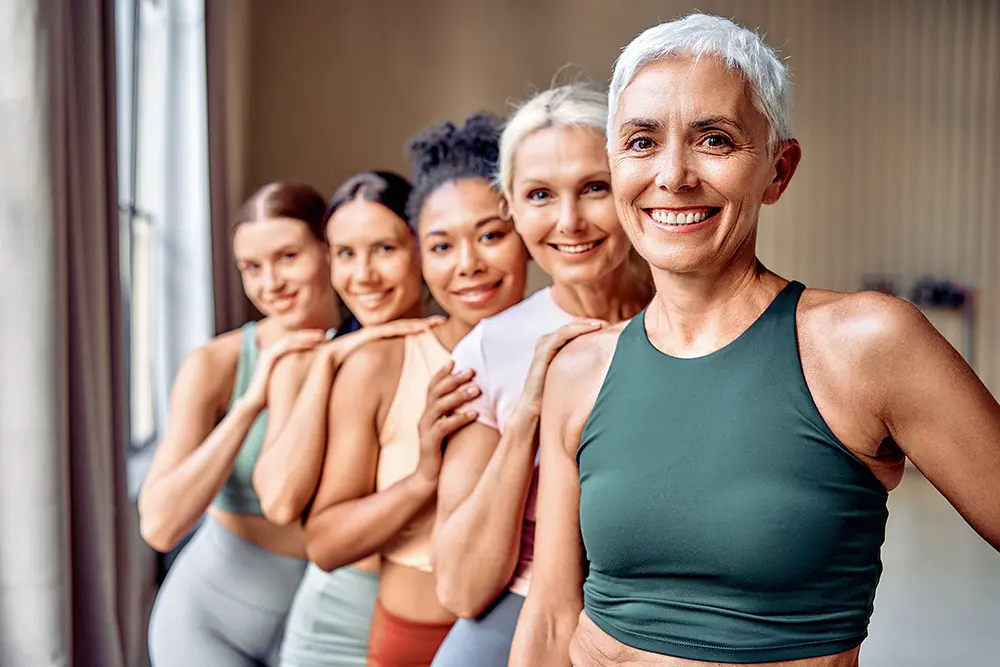 Five women of diverse ages and backgrounds stand in a row indoors, smiling and resting their chins on their hands. They are wearing athletic wear, suggesting a fitness or yoga class.