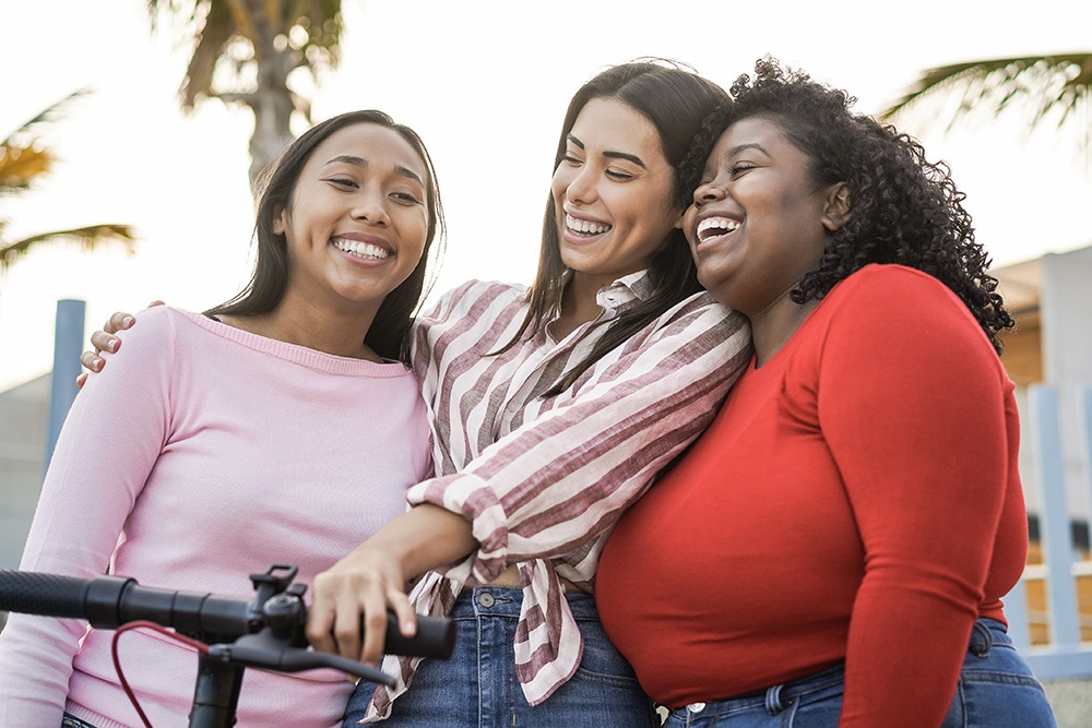 Three women standing outdoors smiling and embracing each other, with palm trees in the background. One woman holds a scooter. They appear happy and are enjoying a sunny day together.