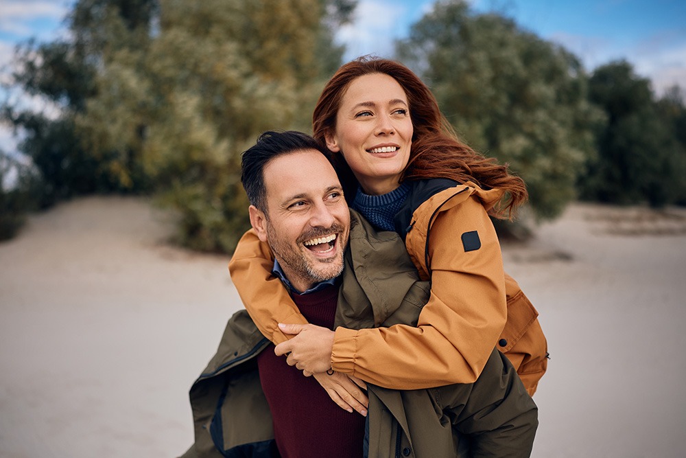 A happy couple embraces outdoors in autumn; the woman leans over the man's shoulder and both are smiling warmly.