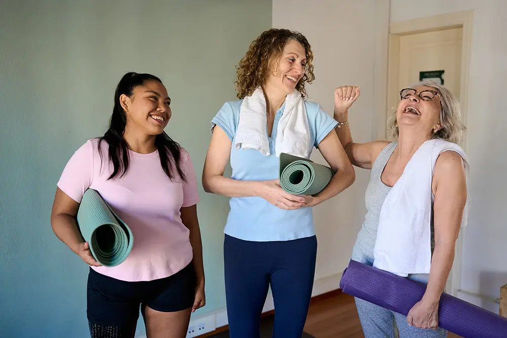 Three women in workout clothes laugh together indoors, each holding a yoga mat and wearing a towel around their neck. They appear happy and relaxed, standing in a well-lit, homey room with light-colored walls.