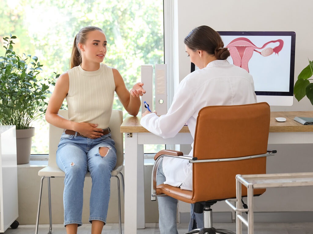 A woman sits holding her abdomen while talking to a doctor at a desk. A computer screen behind them displays a diagram of the female reproductive system.