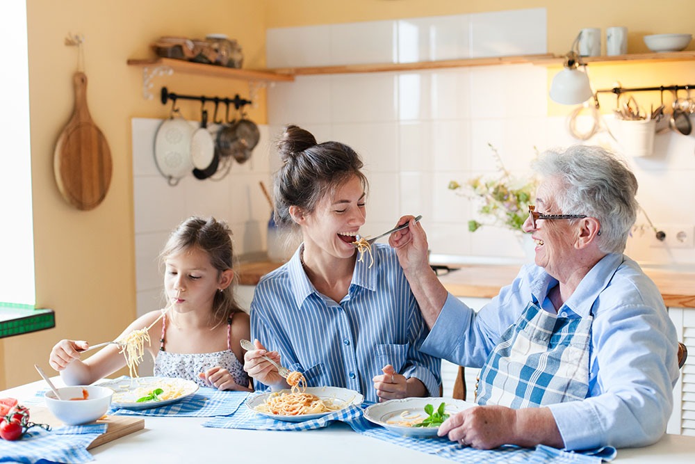 Three generations of women—child, mother, and grandmother—sit together at a kitchen table, smiling and enjoying a meal of pasta. The grandmother is playfully feeding the mother with a fork.