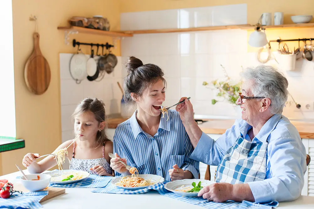 Three generations of women—child, mother, and grandmother—sit together at a kitchen table, smiling and enjoying a meal of pasta. The grandmother is playfully feeding the mother with a fork.