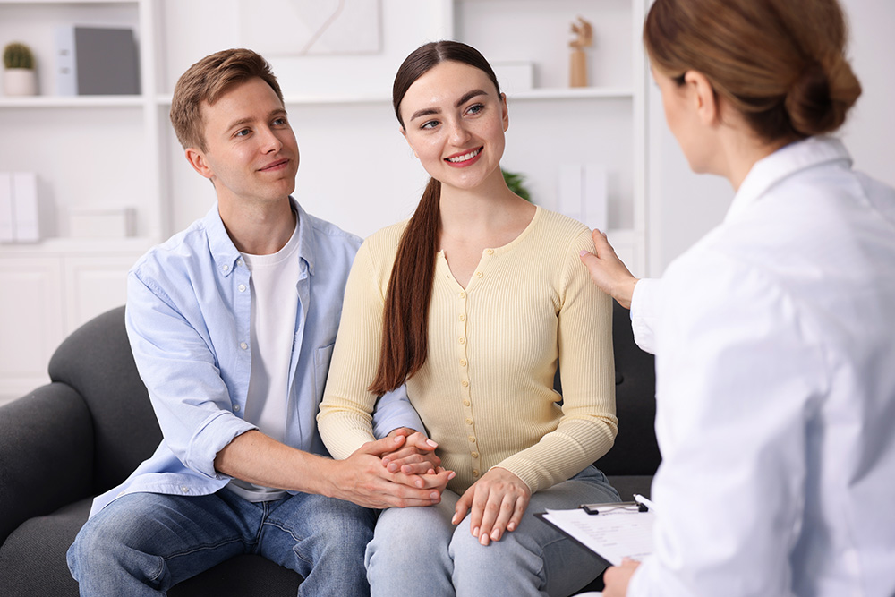 A couple sits on a couch, holding hands and smiling, while a doctor or counselor, partially visible, talks to them and touches the womans shoulder reassuringly.